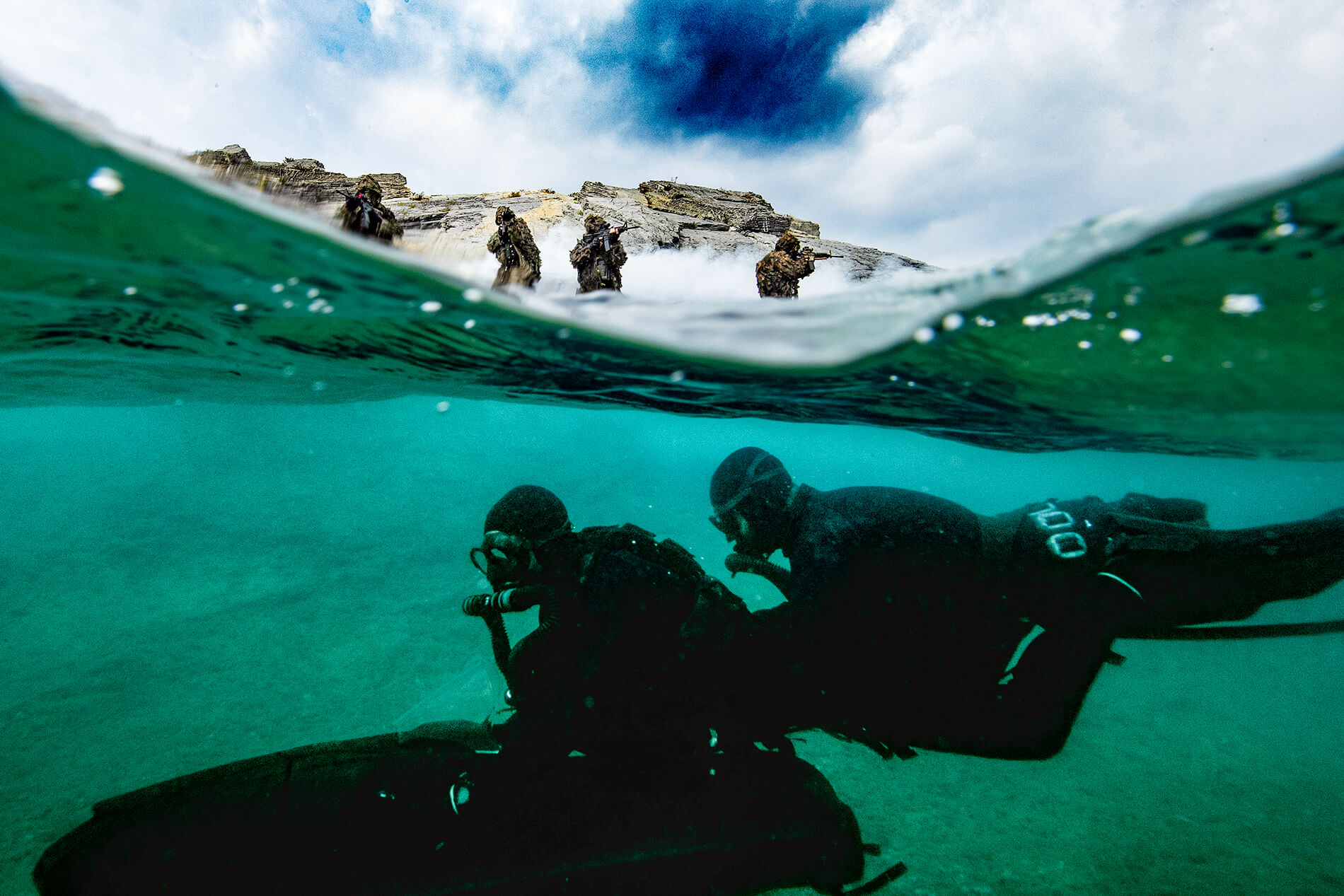 Due incursori della Marina in immersione con trascinatore subacqueo, in superficie 4 incursori che controllano la presenza di minacce